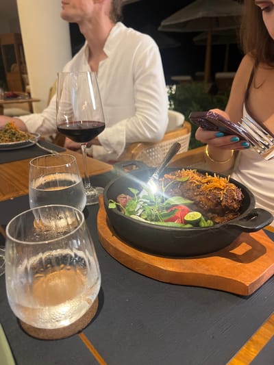 In Tegallalang, Indonesia, two individuals are seated at a table outdoors at night, engaged in a meal. On the table, a glass of red wine, a glass of iced water, and a serving dish containing a meat entree are visible. The entree, presented in a black cast-iron pan, appears to be a piece of meat topped with fried shallots and garnished with fresh herbs, a sliced tomato, and a lime wedge. A fork and knife are placed on the table near the entree. The person on the right is holding a smartphone and a fork. The individual on the left is wearing a white shirt. The background suggests an open-air dining setting with umbrellas and foliage.