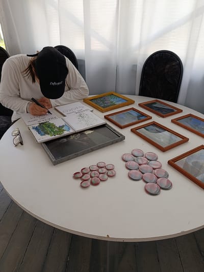 A person wearing a white long-sleeved shirt and a black baseball cap is sitting at a round white table, drawing in an open notebook. The notebook lies on top of a dark gray rectangular object. To the left of the notebook are a pair of eyeglasses. Several framed pictures with wooden frames are arranged on the table, some containing artwork. In the foreground, to the right, are two clusters of oval-shaped, translucent objects with red and green coloring. The scene appears to be indoors, with sheer white curtains in the background and a dark wooden floor beneath the table. The text on the person's cap is not fully legible but appears to include the letters "EXNMOST." The writing in the notebook is in Cyrillic script. The image was taken in Chaykovsky, Russia.