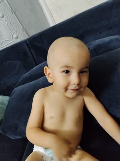A bald toddler, wearing a diaper, rests on a dark blue couch. The child has a fair complexion, dark eyes, and a slight smile. The scene appears to be indoors, with a textured surface visible in the upper left corner, possibly a wall or furniture. The lighting is soft, suggesting it could be daytime. The overall mood is relaxed and domestic. There is no visible text in the image.