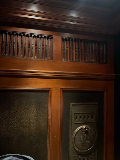 The image depicts the interior of an elevator, likely in a building in New York City, United States of America. The elevator car features ornate, dark wood paneling. Above the main paneling, there are two rectangular decorative grilles with vertical slats. The lower section of the elevator car is divided into two panels. The left panel is covered in a textured dark material, possibly leather or a similar fabric. The right panel features an antique elevator control panel. This panel is made of dark metal and has multiple toggle switches arranged horizontally in two rows. Centered within this panel is a circular plaque with embossed text that reads "GURNEY ELEVATOR" and below that, "GENERAL ELECTRIC CONTROL." A small, circular brass fixture, possibly a speaker or indicator light, is positioned above the text on the plaque. The overall aesthetic suggests an older, possibly historic, building.