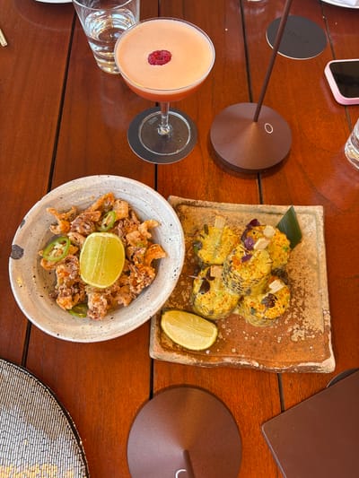 The image shows a wooden table at what appears to be a restaurant in Cannes, France, based on the objects in the scene and the implication that the photo was taken from a patron's perspective. On the table, there is a cocktail in a stemmed glass, adorned with a raspberry on top. Adjacent to the glass are drinking glasses. A plate of what appears to be elote-style corn is also on the table, garnished with small leaves, and a lemon wedge. A bowl of fried calamari or a similar dish, garnished with jalapeno slices and a lime wedge, is placed nearby. Other items visible on the table include a small, dark bronze colored lamp, a cell phone, and a placemat with an intricate woven pattern. The scene suggests a casual dining experience at a restaurant.