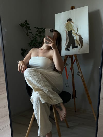 A young woman with long brown hair is taking a mirror selfie in what appears to be an artist's studio. She is wearing a white strapless top and textured cream-colored pants, and is sitting on a black chair. Her right arm is raised, holding a smartphone to capture the reflection. Beside her, an easel holds a partially finished painting of a couple embracing. A potted plant with green leaves is visible in the background, to the left of the woman. The room has wooden flooring and a gray wall. The overall lighting is dim, with light coming from the right side of the frame. The scene does not provide any specific indicators of being located in Ternopil, Ukraine, or any other specific city.