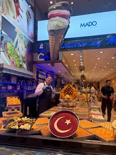 This image depicts the interior of a Mado shop in Beyoğlu, Turkey. The scene is vibrant and displays a wide variety of Turkish desserts, primarily baklava and Turkish delight, arranged in trays and boxes. In the foreground, a man wearing an apron with the Mado logo and blue gloves is giving a thumbs-up to the camera, smiling. He appears to be a staff member. Behind him, a large digital screen displays the Mado logo and Turkish text. Another employee, wearing glasses and a black shirt, is seen in the background on the right, holding a bottle and a pink cloth. Other customers are visible in the distance. The shop is brightly lit with numerous ceiling lights. A large decorative ice cream cone hangs from the ceiling, and a Turkish flag motif is prominently featured on a circular tray of red dessert. The overall atmosphere suggests a busy and appealing retail environment focused on traditional Turkish sweets.
