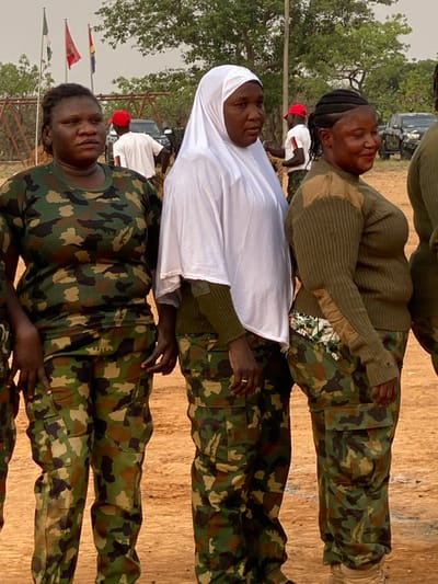 Three women in camouflage uniforms are standing outdoors. The woman in the middle is wearing a white hijab. Flags are visible in the background. The setting appears to be a field or open area with trees in the distance. A few people are partially visible in the background. Vehicles are also present in the distance.
