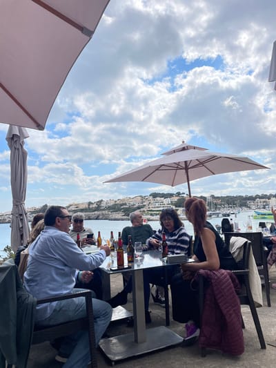 An outdoor scene in Porto Cristo, Spain, depicts a group of six adults seated around a square, reflective table under a partially cloudy sky. The table is laden with multiple amber-colored glass bottles, some containing liquid, and clear drinking glasses. A dark-haired male in a light blue shirt and jeans occupies the left foreground, turned towards the right, with a dark jacket draped over his chair. To his right, a male wearing sunglasses is observed looking at a smartphone. Further right, another male with grey hair in a dark green sweater gazes towards the water. Opposite him, a female with dark, curly hair in a horizontal blue and white striped top is engaged in conversation, gesturing with her left hand. To her right, a female with reddish-brown hair, wearing a black sleeveless top, is also engaged in conversation, with a maroon quilted jacket draped over her chair. Two large light-colored patio umbrellas provide shade, one partially visible in the upper left, and another, fully open and tiered, positioned centrally. In the background, a tranquil bay with blue water is visible, lined with numerous white and light-colored buildings along the shoreline and on distant hillsides. Several boats are moored in the harbor to the right. Green foliage is scattered throughout the landscape.