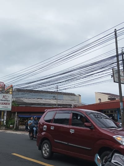The image depicts a street scene in the city of Pacet, Indonesia, under an overcast sky. The road in the foreground is asphalt, with a faded yellow dividing line partially visible on the left.
A dark red multi-purpose vehicle (MPV) occupies the mid-ground, moving from right to left. A male driver is discernible through the front driver's side window. Directly behind and slightly to the left of the MPV, a dark-colored scooter is in motion, carrying two individuals. The driver wears a dark jacket, and the passenger wears a light blue or grey top. A portion of another motorcycle, black with chrome elements, is visible in the extreme bottom right corner, positioned behind the MPV.
On the left side of the street, a vertical sign for "Rumah Makan Kalimantan" (Kalimantan Restaurant) with "CIPANAS" printed below is visible. Adjacent to this sign, greenery lines the roadside, preceding buildings. The primary building on the left features a corrugated metal roof and a reddish-brown facade below, likely a commercial establishment. Further to the right, another building with a similar reddish-brown facade and a contrasting light-colored roof is visible.
A dense array of black overhead electrical or utility wires crisscrosses the upper portion of the frame, originating from the top left and extending diagonally towards the mid-right. A concrete utility pole stands on the right side, supporting these wires and a partially obscured sign. In the background on the right, a building with a sloping light-colored wall and a brown roof is present. The overall lighting is diffuse, characteristic of an overcast day.