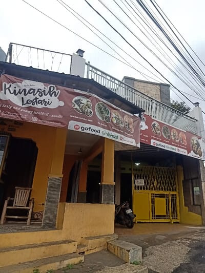 An outdoor, eye-level shot captures the exterior of a two-story building, likely a restaurant, under an overcast sky. The building features a yellow-painted facade with dark grey textured pillars. Concrete steps lead up to the entrance on the left, where a wooden slatted chair rests. To the right, a yellow metal gate and fence secure an entrance, with a dark motorcycle parked in front of it.

A large banner stretches across the front of the building, advertising the establishment. The banner is red-brown with white text and images of various food items, primarily noodle and meatball dishes. Visible text on the banner includes the restaurant's name, "Kinasih Lestari," and a slogan "a tasty and yummy meatball - noodle." An address is also present: "Jl. Cipageran No. 67 Cimahi Utara - 40511," along with a social media handle "baksayaminkinasih." Several food delivery platform logos are prominently displayed, including "GoFood," "GrabFood," and "ShopeeFood," indicating delivery services are available. One menu item explicitly mentioned is "Mie/Bihun Bakso Kuah."

Overhead, numerous power lines crisscross the frame against the pale sky. The upper floor of the building has a white railing, and a stone-like structure is visible behind it. The overall impression is a bustling local eatery in an urban or semi-urban setting.