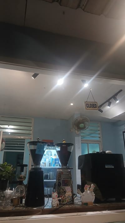Here is a detailed description of the image:
The image appears to be taken from inside a business, likely a coffee shop or café, looking out through a window. The photo is angled so that the right side shows a countertop display, and the left shows the exterior view through the window.
Inside the shop, there are two coffee grinders, one stacked above the other, on the countertop. Both grinders are black, conical shaped. The grinder on top has several stickers attached to its side, and various figurines are arranged around them. Behind the coffee grinders, there's an Espresso machine and a refrigerated display case.
Through the window, a ceiling fan is visible, along with a "CLOSED" sign hanging near a light fixture and track lighting. The background suggests an interior space with a window. The style of the window and the ceiling fan hint at a warm climate.
Based on the presence of "Kecamatan Pati, Indonesia" in the metadata, the location could be identified. However, without further information about the specific business or building, it's difficult to determine the exact location. The style of the interior and the appliances are consistent with a coffee shop in a Southeast Asian urban area.