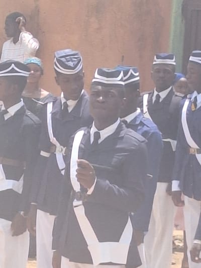 A group of men are standing in a row, wearing dark-colored uniforms with white trim and hats with a white design. They are in Sabon Tasha, Nigeria, holding what appear to be ceremonial swords. One person is visible to the left of the group and appears to be wearing a light-colored shirt. The background appears to be a dusty, outdoor area.