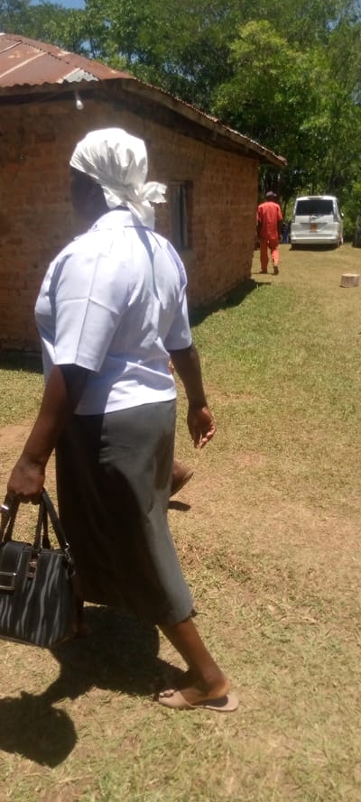 The image shows a woman walking away from the camera, carrying a black handbag. She is wearing a white shirt and a white headscarf. The woman's right foot is visible, she is wearing sandals. There is a brick building in the background, to the left of the image. There are trees in the background to the right of the image. A man in an orange shirt is walking away from the camera, to the right of the image. He is in the distance, walking on a paved road. There is a white car parked on the side of the road, to the right of the image. There is a patch of grass on the right side of the image. The coordinates 0.537, 34.799 place this image in Lwandeti, Kenya.