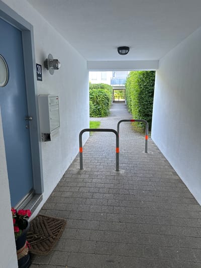 The image depicts an outdoor hallway or passage in Höhenkirchen-Siegertsbrunn, Germany. The hallway has white walls and a white ceiling. The floor is paved with gray rectangular stones. On the left side, there is a light blue door with a round window and a silver doorknob. Next to the door, on the wall, is a black sign with the number "38" in white and the text "Lerchstr." below it. Below the sign is a white mailbox. Further down the hallway, on the left, a pot with red flowers sits on the ground next to a brown doormat. In the center of the hallway, there are two grey metal bicycle racks with orange reflective bands near their base. The hallway is lined with neatly trimmed green hedges on both sides, which continue to the end of the passage, where daylight and buildings are visible in the distance. A black light fixture is mounted on the ceiling in the middle of the hallway. Another light fixture, with a metal cage around the bulb, is mounted on the wall to the left, near the door and sign. The ground appears to be slightly damp.