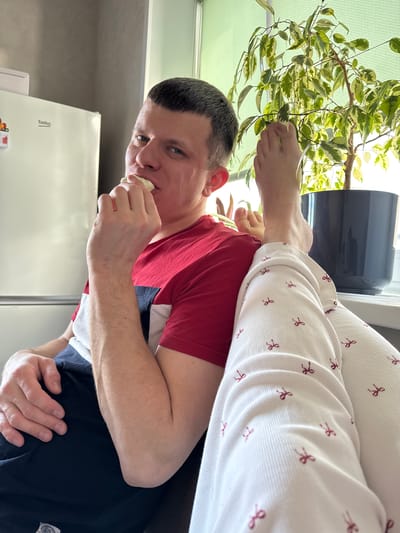 A medium-shot photograph taken in a domestic interior in Gornyi (Dvurechenskoe S/P), Russia, depicts an adult male and the lower extremities of a second individual. The male subject, with short dark hair and blue eyes, wears a red, white, and navy striped t-shirt and navy shorts. He is seated, looking directly at the camera with a slight smirk, and holds a light-colored food item to his mouth with his right hand. His left arm rests on his lap. Bare feet and lower legs of a second individual, clad in white ribbed pants with a red bow pattern, rest across the male subject's lap/thigh. In the background, a white Beko refrigerator with a magnet on its door is partially visible against a dark grey wall. To the right, a potted variegated Ficus plant sits on a windowsill, illuminated by natural light entering through a window covered by a light green roller blind.