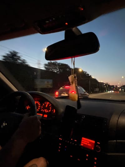 The image is taken from inside a car, looking forward, at dusk or dawn. The driver's hand is visible on the steering wheel, and the dashboard lights are illuminated red. A car air freshener hangs from the rearview mirror. Outside, traffic lights and streetlights are visible, along with trees and buildings. The sky is a gradient of blue and orange. The location is identified as Velyki Hai, Ukraine.