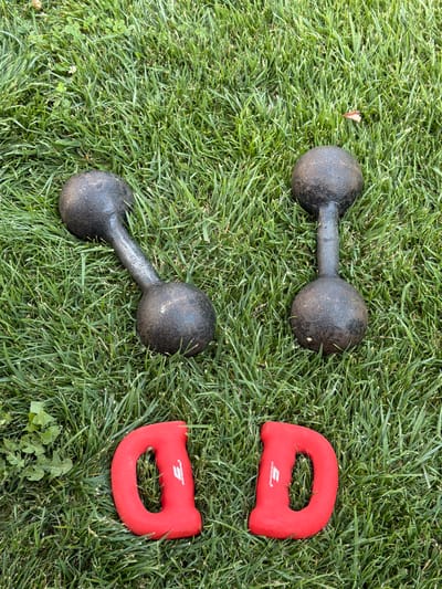 Two black dumbbells are placed on a grassy lawn. In front of the dumbbells, two red hand grip strengtheners are arranged to form the letter "D". The scene is outdoors, likely in a park or backyard, with lush green grass. No people are present in the image. The overall setting does not provide specific clues about the location within the city of Cholpon-Ata, Kyrgyzstan.
