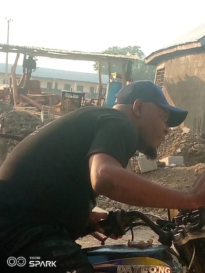 The image portrays a male individual situated on a motorcycle in an outdoor environment within the city of Ajaokuta, Nigeria. The man, viewed from his right side, wears a dark blue baseball cap and a black short-sleeved t-shirt. He has a beard. His right hand grips the motorcycle's handlebar. The visible portion of the motorcycle includes a dark blue fuel tank with a gold stripe and the word "DAYLONG" inscribed on it.
The background depicts an area consistent with construction or undeveloped land, characterized by extensive piles of dirt, gravel, and scattered debris including concrete blocks. In the mid-ground, a dilapidated or unfinished shelter with a corrugated metal roof supported by wooden posts stands before a lighter-colored building with a dark roof. To the right, a separate building constructed of light brown material with a dark roof is visible. Daylight illuminates the scene, casting shadows and contributing to a visible atmospheric haze.