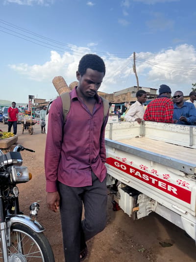The media file is a portrait of a man in Rafin Par, Nigeria. The man is wearing a purple button-up shirt and dark pants, with a woven basket slung over his shoulder. He is standing in front of a white truck with red lettering that reads "GO FASTER" in English and Chinese characters. The truck appears to be carrying several passengers in its bed.
The scene suggests a market or a busy roadside area. In the background, there are buildings, a power pole with wires, and other vehicles, including a motorcycle in the foreground. The weather appears to be sunny with some clouds in the sky. The time of day seems to be daytime, likely late morning or early afternoon. The man's expression is neutral, and the overall mood of the image is that of everyday life in a bustling environment. The presence of the truck with people indicates transportation and community activity. The man's attire and the basket suggest he might be a vendor or a traveler carrying goods. The setting, with its unpaved ground and simple structures, points to a rural or semi-urban area.