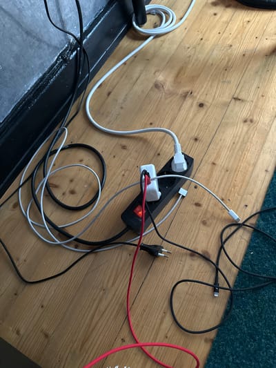 The image shows a close-up, overhead view of a power strip with several cables plugged into it, lying on a wooden floor. The power strip is black with a red on/off switch that is illuminated, indicating it is on. A white cable is plugged into the top socket, and a white USB adapter is plugged into another socket, with a short white cable connected to it. There is also a small white adapter plugged into one of the sockets. A red and black cable is also connected to the power strip. Several other black and white cables are coiled or spread across the wooden floor, some leading away from the power strip. A section of a dark wall or furniture is visible in the upper left corner, and a textured green surface, possibly carpet or a rug, is visible on the right side. The scene appears to be indoors, likely in a residential or office setting. No specific landmarks or indicators of Hamburg, Germany, are visible in the image.