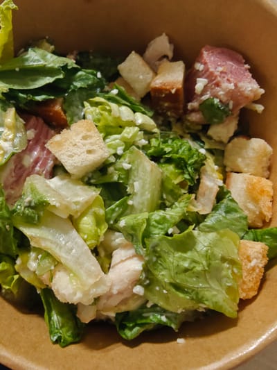 The image shows a close-up, overhead view of a Caesar salad in a cardboard bowl. The salad contains chopped romaine lettuce, croutons, and chunks of what appears to be ham and cooked chicken, all coated in a creamy dressing. There are also flecks of white cheese visible throughout the salad. The salad is presented in a light brown, textured cardboard container, suggesting it might be takeout or a casual dining setting. The lighting is dim, and the focus is on the food, with the background slightly blurred. The image does not contain any information about a specific location in Dubai or the United Arab Emirates.