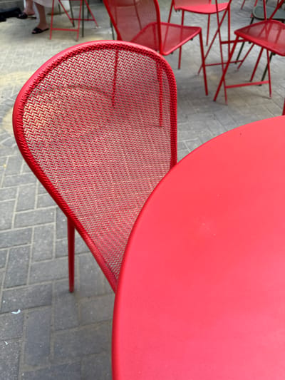 The image depicts an outdoor seating area with multiple red metal chairs and a red circular table. The chairs have a mesh design for their backs and seats. The seating area is paved with gray rectangular bricks. In the background, the legs of people can be seen, and some of them are wearing black flip-flops. The scene appears to be a casual outdoor cafe or patio. The color palette is dominated by red and gray. There are no specific landmarks or architectural details to pinpoint the location to Riga, Latvia, or any other specific city.