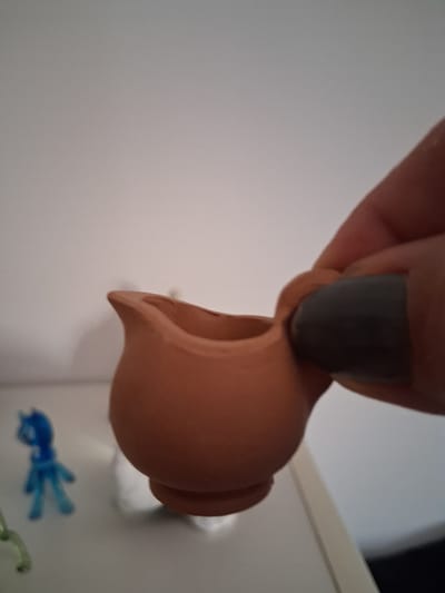 A person's hand, with dark grey nail polish, is holding a small, terracotta-colored jug. The jug is round with a flared rim and a short neck. The hand is positioned above the jug, and it appears the person is either holding it for display or is about to pour something from it. In the background, a white surface is visible, possibly a shelf or a wall. There are also some blurry blue objects in the lower right corner, which appear to be small plastic toys. The lighting is soft and diffused, suggesting an indoor setting. No text is visible in the image. The overall scene is simple and focuses on the small jug and the hand holding it.