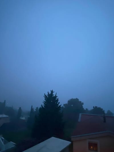 The image captures a moody, misty, or foggy scene at dusk or night in Šušanj, Montenegro. The sky, which occupies a significant portion of the upper frame, is a uniform deep blue-grey, suggesting either heavy cloud cover, twilight, or thick atmospheric conditions.

Silhouetted against this blue expanse, primarily on the right side of the frame, are dense, dark forms of trees, likely conifers given their pointed shapes. To the right, a portion of a building with a reddish roof and light-colored walls is visible. A single window in this building glows with a warm, yellowish light, contrasting with the cool, dark tones of the exterior scene. Below and around the main cluster of trees and the visible building, indistinct shapes of other structures and vegetation are shrouded in the pervasive mist or darkness, extending towards the bottom and left edges of the frame.

There is no discernible activity taking place; the scene is static and quiet. The notable details include the atmospheric blue hue dominating the scene, the stark silhouettes of the trees, and the solitary illuminated window, which provides a small point of warmth and human presence within the otherwise dark and natural setting. No visible text is present in the image.