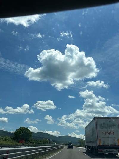 The image depicts a view from a vehicle traveling on a highway in Verinsko, Bulgaria, under a bright, cloudy sky. A large, white truck with the company name "HNR" and website "www.hnrlogistics.com" is prominent in the foreground, to the right. A dark-colored car is visible ahead on the highway, to the left of the truck. The highway is bordered by guardrails and a grassy verge. In the background, lush green hills covered in trees rise under a sky filled with fluffy white cumulus clouds and wispy cirrus clouds. The overall scene suggests a sunny day on a road trip through a scenic, rural area.