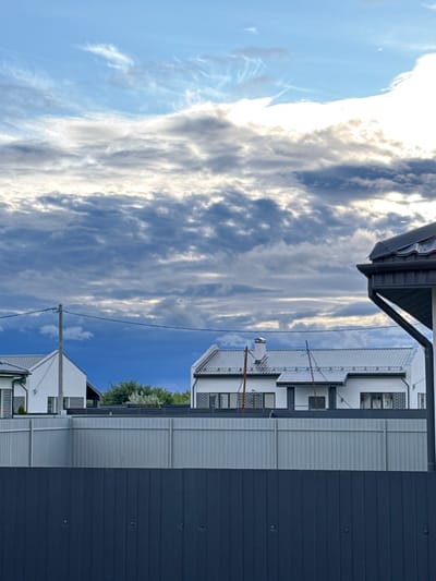 The image depicts a view looking upwards at a building with a dark blue corrugated metal exterior on the right and white sections with grey ribbed siding on the left. The building has multiple levels and windows. Above the building, a bright sky is visible with a mix of blue sky and dramatic, elongated white and grey clouds, suggesting it is daytime. Sunlight is breaking through the clouds, illuminating some of them brightly. The perspective is from a low angle, possibly looking up from street level. There are hints of greenery from trees visible behind the building in the distance. A utility pole with wires is also present in the frame, extending upwards into the sky. The overall scene is urban or suburban.