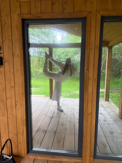 An eye-level shot captures a woman striking a dynamic pose outside a modern wooden cabin, viewed through a large rectangular window. The image is oriented vertically, with the cabin's interior walls made of horizontal wooden planks framing the window. The window itself has a dark, possibly grey or black, frame.

The woman, seen through the clear glass, is wearing light-colored, likely white or off-white, casual attire consisting of a long-sleeved top and wide-leg trousers. She has long hair and is holding a smartphone, seemingly taking a selfie. Her body is angled, with one leg bent and slightly raised, and her arms are up, one holding the phone. She appears to be standing on a wooden deck or porch outside the cabin.

Beyond the deck and the woman, the outdoor scene reveals lush green foliage, possibly grass or bushes, under bright daylight. The overall setting suggests a natural, possibly remote or vacation, location, consistent with the provided context of Vozhoiskii, Russia. On the interior floor of the cabin, in the bottom right corner, a black shoulder bag or purse is visible. The bright lighting suggests it is daytime. No discernible text is visible in the image.