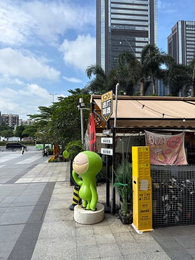 In the Haizhu District of China, a paved walkway stretches past a light green, cartoonish alien sculpture standing on a white pedestal. Behind the sculpture, a yellow utility box is stacked vertically. To the right of the utility box, a wire mesh enclosure contains bottles and other refuse. A beige awning extends over an outdoor seating area of a establishment. A banner hangs from the awning, displaying the word "Beer" and an illustration of a beer mug. A street sign with Chinese characters and the number "520" is mounted on a pole near the sculpture. In the background, tall, modern buildings rise into a blue sky with scattered clouds. Palm trees and other greenery are visible between the buildings and the walkway. A black van drives on a street in the distance, and several people are walking on the walkway.