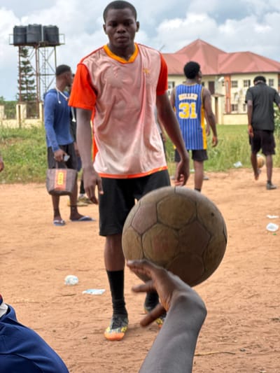 A vertically oriented, candid shot captures a group of young Black men in an outdoor, unpaved area, likely in Ihiagwa, Nigeria, during the daytime under an overcast sky.
In the foreground, a young man wearing a light orange/peach sleeveless shirt with orange trim and dark shorts looks directly at the camera with a neutral expression. His arms are bent, suggesting a paused motion or relaxed stance. To the right, a weathered, light brown soccer ball is held by an unseen person's hand, positioned close to the camera, indicating an immediate involvement in a game or sports activity.
Several other young men are visible in the mid-ground and background, some blurred due to motion or shallow depth of field. One man behind the main subject wears a blue and yellow jersey with the number "13" prominently displayed on the back. Another person in the background wears a black shirt and shorts, while a third is seen in a blue top. On the ground, scattered small pieces of white trash are visible, and a pair of light-colored flip-flops are worn by a person in the mid-ground, who also has a small brown tag or sticker on their shorts with partially visible text that appears to be "DOGN" or similar.
The setting includes patches of green grass, reddish-brown earth, and buildings in the background, one with a distinctive reddish roof and another with multiple windows. To the far left, a tall metal structure supports several large black water tanks, a common utility fixture. The overall scene suggests a casual gathering or a game of soccer (football) among friends.