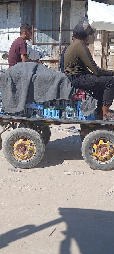 A side view of a cart loaded with beverages, likely sodas or energy drinks in blue cans, is shown. The cart is pulled by a vehicle which is not visible. Three men are seated on the cart. The man in the foreground on the right is wearing a dark green long-sleeved shirt, black pants, and sandals. The man in the middle is wearing a dark blue long-sleeved shirt and a baseball cap. The man on the left is wearing a maroon t-shirt. A gray blanket is draped over some of the cargo. The cart has two wheels visible, one in the foreground and one behind it. The wheels are old and worn, with yellow rims. The background shows a weathered metal structure, possibly a makeshift stall or building, and some construction materials. The ground is dirt and sand. The scene appears to be an outdoor market or street in Khan Yunis, Palestinian Territories.