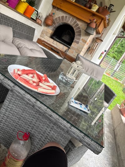 The image shows an outdoor patio setting, possibly in a backyard. A glass-topped table with a grey wicker frame is the central focus. On the table, there is a white oblong plate filled with imitation crab sticks. There is also a lantern, a clear glass, and a phone case. Under the table, there is an empty plastic bottle and a person's leg. In the background, there is an outdoor fireplace with decorations, a wicker sofa with cushions, and a view of a garden through a fence. The image was not necessarily taken in Pernik, Bulgaria.
