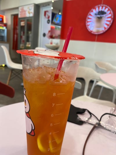 A close-up shot of a clear plastic cup filled with a bright orange liquid. The cup is labeled "O TEA" and has a red lid with a cartoon image of a snowman. A red straw sticks out of the top of the cup. The liquid in the cup is partially filled with ice, and the level is marked with measurements. The cup is sitting on a white table. The table is partially out of focus, but there are glasses, a black face mask, and a white chair in the background. The environment is a cafe or restaurant with red and white walls. There is a large red circle with a white interior on the wall behind the cup. The location is a shop in Kecamatan Piyungan, Indonesia, latitude: -7.837, longitude: 110.474.