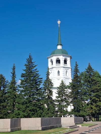 The image depicts a view of the Epiphany Cathedral in Irkutsk, Russia, on a clear day. The cathedral's white bell tower, topped with a green spire and a golden cross, stands prominently against a bright blue sky. A line of tall, dark green pine trees partially obscures the lower sections of the tower, creating a visual contrast between the man-made structure and the natural elements. In the foreground, a low, rectangular wall made of stone blocks and a patch of green grass are visible. The scene is devoid of people.