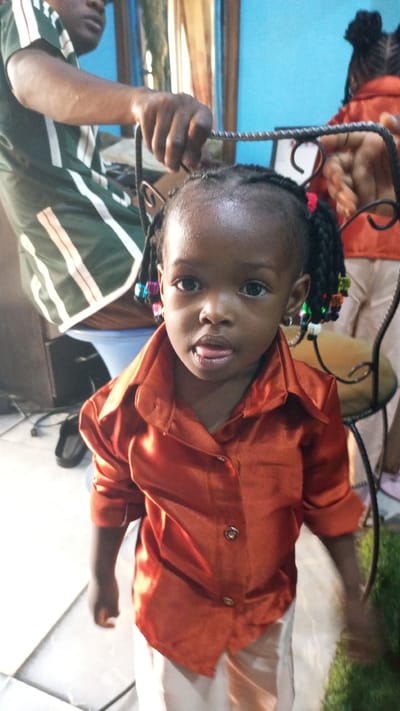 A young, dark-skinned child stands indoors, wearing a shiny, orange button-down shirt and light-colored pants. The child has braided hair adorned with colorful beads. Behind the child are two partially visible adults, one in a green and white shirt and another in a matching orange shirt. There is a metal object held near the child's head. The location is presumed to be Sabon Tasha, Nigeria.