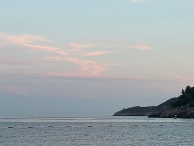 This is a horizontal, eye-level, medium shot of the Adriatic Sea at dusk near Bečići, Montenegro. The sky is a gradient of soft blues and pinks, with wispy pink clouds streaking across the upper portion of the frame. The sea is calm, with gentle ripples reflecting the sky. A line of buoys, alternating red and white, demarcates a swimming area in the foreground. To the right, a rocky, tree-covered peninsula juts into the sea. A small, square tower is visible on the crest of the peninsula. The overall mood is serene and tranquil.