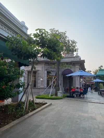The image depicts an outdoor seating area of a establishment, likely a cafe or restaurant, with people seated at tables. The scene is viewed from a slightly elevated angle, looking towards a building with classical architectural elements. In the foreground, a large tree with green foliage partially obscures the view.
Several people are visible, seated at tables in the outdoor area. Some are engaged in conversation, while others appear to be looking around. The people are casually dressed. Objects present include tables and chairs, and two prominent blue and white striped umbrellas, providing shade. The building behind the seating area has stone cladding and large windows. A paved walkway runs alongside the seating area and continues into the distance. There are also planters with greenery.
The time of day appears to be late afternoon or early evening, given the soft, diffused light and the clear sky. The weather seems pleasant. The architecture of the building and the presence of outdoor seating suggest an urban or semi-urban commercial setting. The overall mood is relaxed and social. No visible text is present in the image.