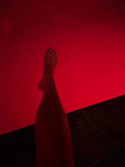 This media file features a close-up shot of a person's lower leg and foot against a vibrant red background. The leg and foot are illuminated by a red light, giving them a reddish hue. The background is predominantly a deep red, with a textured dark area on the right side of the frame. The leg is positioned diagonally, with the foot extending towards the left side of the image. The toes are visible and appear to be relaxed. The scene is abstract and minimalistic, focusing on the form and color. There is no discernible activity or event taking place, and no text is visible. The lighting suggests an indoor setting, possibly a studio or a room with specialized lighting. The location context of Nusa Penida, Indonesia, does not provide any direct visual cues within the image itself, as the image is abstract and devoid of specific environmental details. The overall mood is serene and perhaps a bit enigmatic due to the dominant red color and the solitary subject.