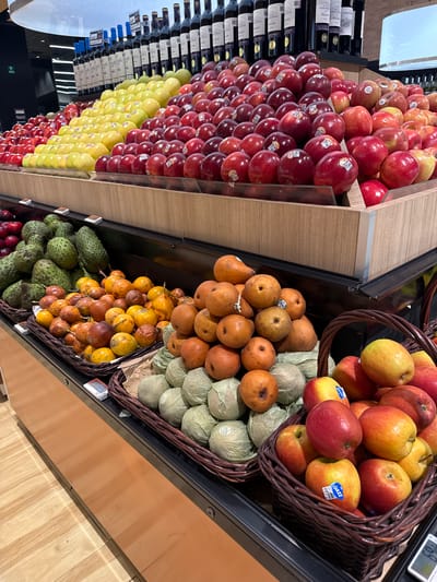 The image shows a grocery store display, likely inside a supermarket, featuring fresh produce and wine bottles. The location is plausibly in Puebla, Mexico, based on the types of fruits and the overall aesthetic.
On the left, stacked apples of different varieties (red, yellow, and a mix) form a large display. Behind the apples, wine bottles are arranged on shelves.
To the right, produce is arranged in baskets on shelves. From top to bottom, the baskets contain:
-A mix of red and yellow apples.
-Orange-skinned pears atop a layer of wrapped pale green fruits (possibly Asian pears or a type of melon).
-Yellow passion fruits.
-Green soursops at the very bottom, mixed with some red fruit.
The shelving and flooring are made of light-colored wood. The lighting is bright, suggesting an indoor supermarket environment. There are no people visible in the frame.