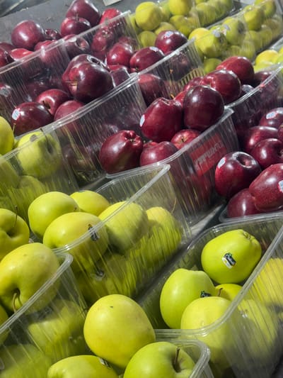 This image captures a close-up, angled view of various apples displayed for sale in what appears to be a retail setting, likely a grocery store or market in Ramat Gan, Israel. The scene features rows of apples, clearly divided into two main varieties: deep red apples and bright green apples.

The apples are neatly arranged in clear plastic trays or containers, stacked or lined up on shelves. The red apples, which are a darker hue, occupy the left and central parts of the frame, while the vibrant green apples are predominantly on the right. Both varieties are numerous, filling their respective packages.

Several of the green apples have visible stickers. One prominent sticker clearly displays "Melinda GOLDEN" along with a small bee illustration, indicating the brand and variety (Golden Delicious). Text on the stickers on the red apples is less distinct, but some appear to be standard produce labels or barcodes. The lighting in the image suggests an indoor environment, possibly from overhead fluorescent lights, creating reflections on the shiny apple skins and plastic packaging. There are no people visible, and no specific time of day or weather conditions can be inferred. The overall impression is one of fresh produce ready for purchase.