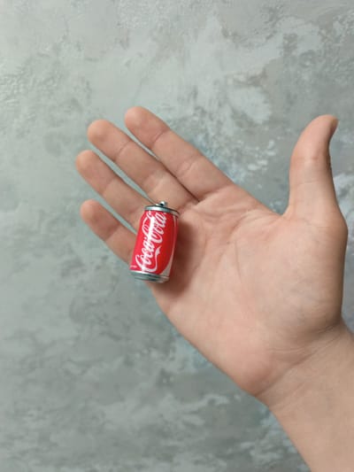A small, red and white miniature Coca-Cola can rests on the open palm of a fair-skinned hand. The can's design is detailed, mimicking a standard Coca-Cola can. The hand is positioned against a mottled grey and white background.