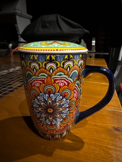 A vibrantly decorated ceramic mug with a lid and an attached tea strainer sits on a wooden table. The mug features a mandala-like pattern in shades of blue, yellow, red, orange, and green. The lid is light green with yellow and red accents. A small orange string dangles from the lid. The background is dark and out of focus, showing what appears to be outdoor furniture or equipment covered by a black tarp, and part of a building structure. The lighting suggests it is evening or nighttime. The image was taken in Heiligenrode, Germany.
