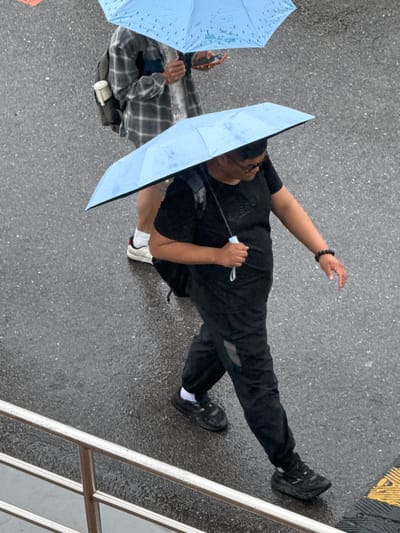 An overhead shot shows two people under blue umbrellas on a wet, grey asphalt surface. One person is wearing all black clothing and black sneakers with white socks, holding their umbrella in one hand and reaching out with the other. The second person is wearing a grey and black plaid shirt and holding a smartphone. A white cup is visible on the ground near the second person. A silver railing is visible in the bottom right corner, and yellow and black markings are visible in the top right corner. The environment suggests a rainy urban setting. The location of Zhongtun, China is not determinable from the image.
