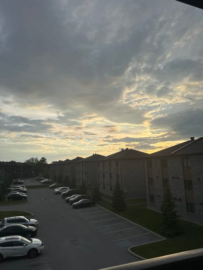 The image depicts a view of apartment buildings and a parking lot, with a dramatic cloudy sky overhead. The buildings are brick structures with dark roofs and visible windows. A row of parked cars, including sedans and SUVs, is arranged in the asphalt parking lot. Evergreen trees are planted along the buildings and surrounding the parking area. The sky is filled with gray and white clouds, with patches of yellow and orange light suggesting the presence of the sun, possibly at sunrise or sunset. The scene appears to be in a residential area. This scene is located in Gatineau, Canada.
