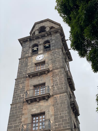 The image displays a tall, imposing stone bell tower, likely part of a church or historic building, viewed from a low angle against a uniformly overcast, light grey sky. The tower is constructed from dark, rectangular stone blocks, giving it a rugged and ancient appearance.
Towards the upper section, a circular clock face with Roman numerals from I to XII is clearly visible. The clock's hands indicate a time of approximately 1:50 or 2:50. Adjacent to the clock face and slightly above it, an arched opening reveals a large bell within, confirming its function as a bell tower. Another bell opening is partially visible further up.
Below the clock and on the lower visible sections of the tower, several rectangular windows are present. These windows are adorned with intricate, black wrought-iron grilles and feature wooden frames, adding decorative detail to the sturdy stone facade. A metal downspout runs vertically down one side of the tower, indicating rainwater drainage. The architecture also includes various decorative cornices and ledges that articulate the tower's levels.
In the upper left portion of the frame, the dark green foliage of a tree partially frames the tower, suggesting the presence of greenery nearby. The sky is completely clouded over, indicating an overcast daytime. No people or specific activities are depicted; the scene is static, focusing solely on the architectural structure.
The only visible text consists of the Roman numerals on the clock face. The architecture is consistent with historical European structures, and the provided context places this location in San Cristóbal De La Laguna, Spain.