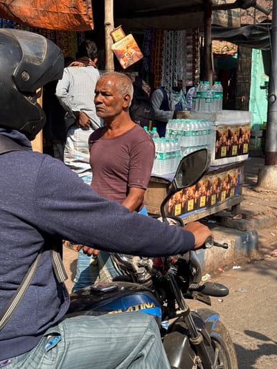A sunlit street scene shows a row of stacked bottled water and snack packages behind a vendor resting. He leans on a striped cushion beside a book. A motorcyclist's arm and bike occupy the foreground. The helmet wearer's blue sweatshirt is prominent. The scene shows daily life and commerce in an urban setting.