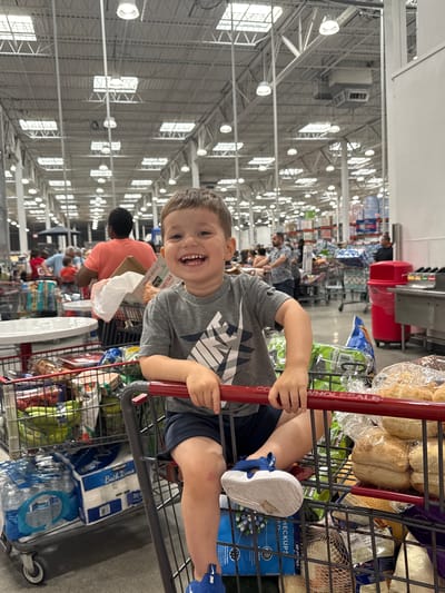 A young boy, appearing to be around 4-5 years old, is seated in a shopping cart filled with groceries. He is wearing a grey t-shirt with a black and white graphic design and dark blue shorts. He has a wide, joyful smile and is looking towards the camera. His right hand is resting on the handle of the shopping cart. The shopping cart is stocked with various items including bottles of water, toilet paper, bags of produce, and loaves of bread. The background suggests an indoor retail environment, likely a large warehouse store such as Costco, with high ceilings, overhead lighting, and numerous shelves stocked with merchandise. Other shoppers are visible in the distance, as are more shopping carts. The overall scene is brightly lit. The location is the City of Yonkers, United States of America.