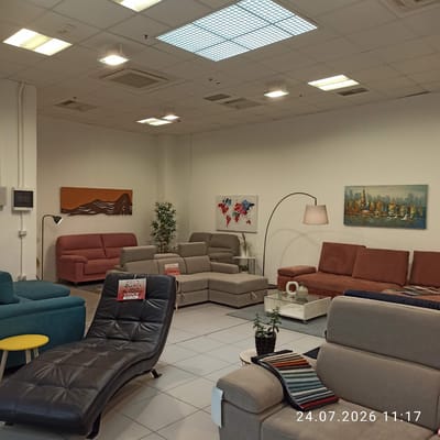 The image shows the interior of a furniture store, likely a showroom, in Modugno, Italy. The showroom is well-lit with overhead fluorescent and grid lighting. Various sofas and furniture pieces are displayed on a tiled floor. In the foreground, a dark leather chaise lounge is prominently featured with a price tag indicating "VERATELLE € 390,00 STOCK PIEZZO UNICO". To the left of the chaise lounge, a teal sofa is partially visible, and a small yellow side table stands nearby.

Further into the showroom, there are several sectional sofas in neutral tones, including a large grey L-shaped sectional. A floor lamp with a beige shade arcs over a coffee table with glass top. Several pieces of wall art are displayed, including a horizontal abstract piece with earth tones and a colorful world map canvas. A cityscape painting hangs on the far wall. Decorative plants and vases add to the showroom's aesthetic. A large section of the ceiling features a grid-like panel, possibly for ventilation or diffused lighting. The overall impression is one of a modern and spacious furniture retail environment. The timestamp "24.07.2026 11:17" is overlaid on the bottom right of the image.