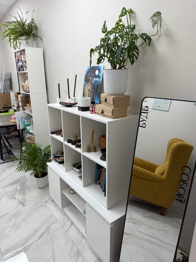 The image depicts an indoor scene, likely a room or studio space, with clean, modern white shelving units and a white tiled floor. Several potted plants are visible, including a large fern on top of a taller shelf unit on the left and a smaller palm-like plant on the floor in front of a lower white unit. On the shelves, various decorative objects are displayed, such as candles, a painting of a cat under a moon, small statues, and stacks of woven boxes. A large standing mirror with a black frame is positioned on the right side of the image, reflecting a yellow armchair and part of the room. Text in Cyrillic is visible on the mirror, reading "БУДЬ СОБОЙ" which translates to "BE YOURSELF". The overall impression is of a well-organized and aesthetically pleasing space. There is no indication of the city of Petropavlovsk-Kamchatsky, Russia, or any specific geographical markers in the image.