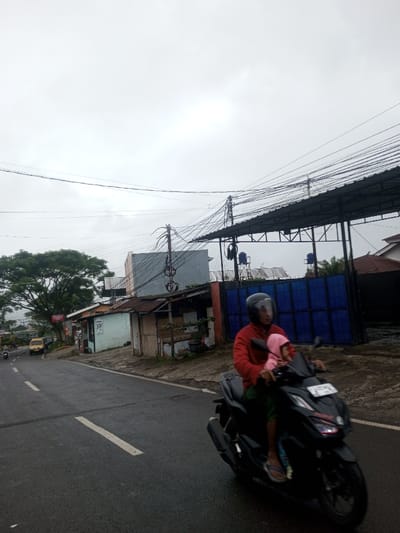This image depicts a street scene in Babaliya Mal., India, under an overcast sky. The asphalt road, appearing wet, features broken white center lines and a solid white line on its right edge.

In the foreground, a dark-colored scooter is in motion, heading slightly rightward. The driver wears a red long-sleeved shirt, dark bottoms, and a black full-face helmet with a clear visor. A child, wearing a pink hooded top and light-colored clothing, is seated in front of the driver, holding the handlebars, and is not wearing a helmet. The scooter has a front license plate, partially obscured.

Along the right side of the road, a dirt shoulder precedes a series of structures. These include several small, utilitarian buildings with corrugated metal roofs and varying wall finishes, such as pale blue paint and unpainted concrete. Further back, a grey concrete building is visible, with a utility pole in front of it heavily adorned with a dense array of overhead power and communication lines. To the far right, a large dark metal canopy extends over a blue gate or wall, behind which two blue cylindrical water tanks are mounted on elevated supports. In the background on the road to the left, another motorcycle and a yellow car are visible.